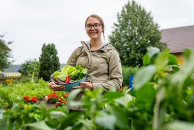 Außen, Tag: Dorfhelferin steht im Garten und trägt eine Schüssel mit Gemüse auf dem Arm.